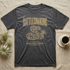 Coiled corn snake mid-tail-vibration in warm tan ink engraving beneath an arched wordmark reading RATTLESNAKING and the subtitle Angry Velcro Mode. — shown on a Dark Heather tee.