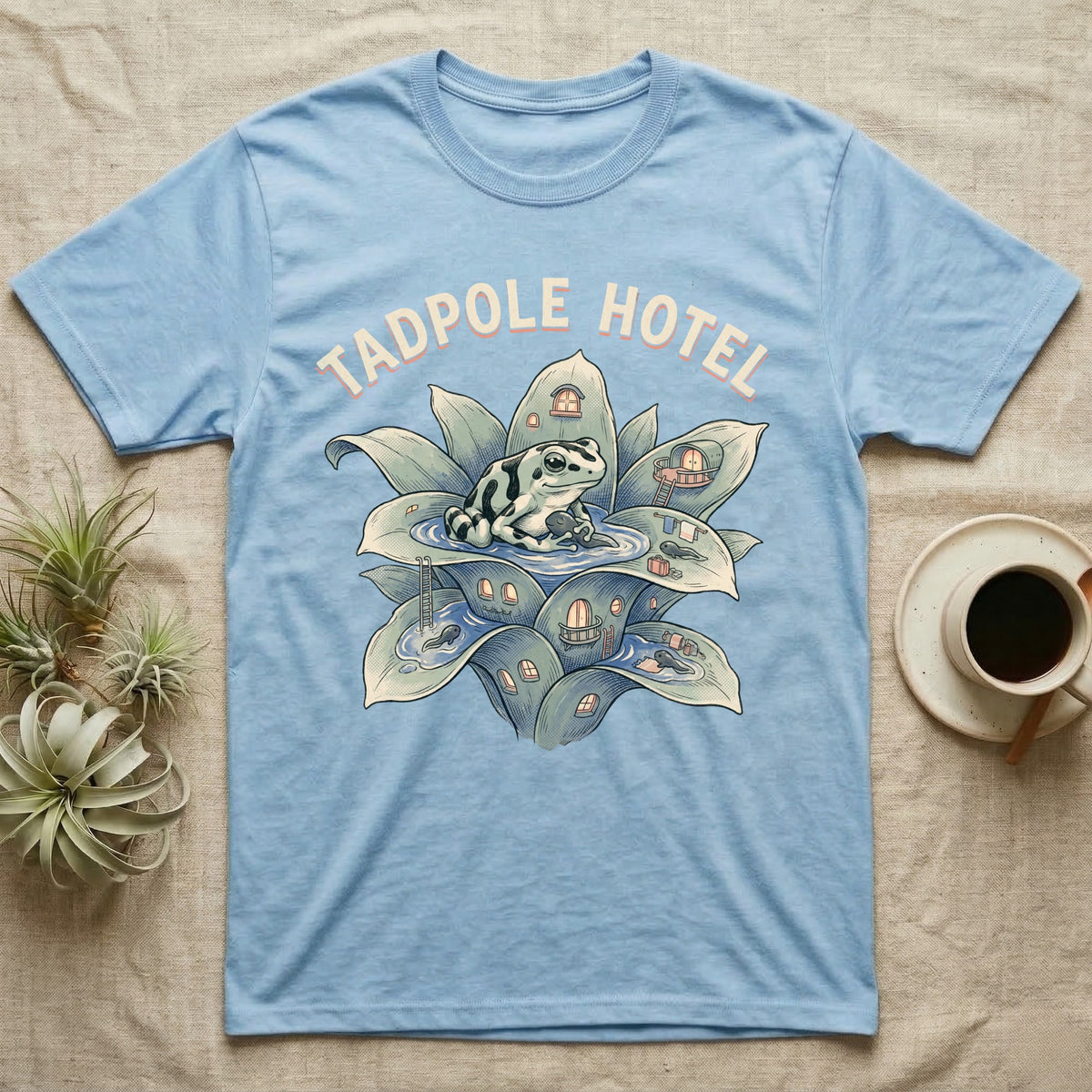 Tadpole Hotel Frog T-Shirt in Light Blue — Whimsical naturalist illustration of a dart frog tucking tadpoles into a bromeliad Tadpole Hotel with hand-lettered marquee text.