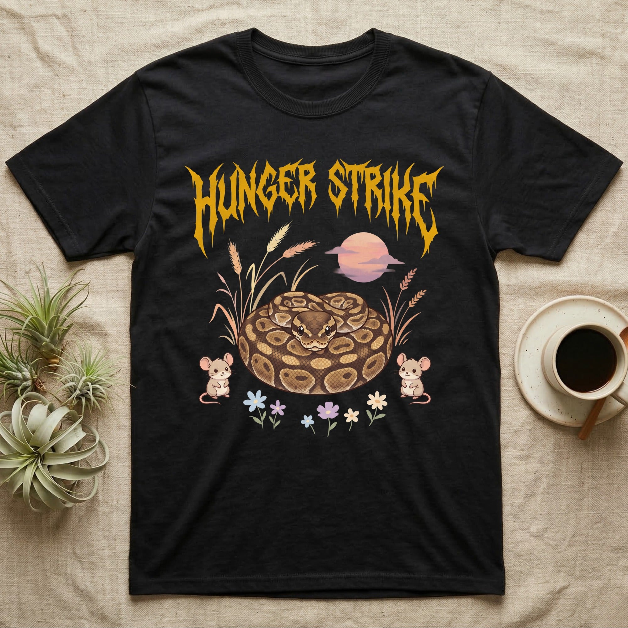 Ball Python Feral T-Shirt in Black — Ball python coiled in a loaf in a pastel savanna scene under a spiky honey-gold metal wordmark reading HUNGER STRIKE.