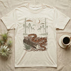 Corn Snake Home Range T-Shirt in Natural — Woodcut-style illustration of a corn snake winding across pine straw beside a split log with pine trees behind, no on-shirt text.