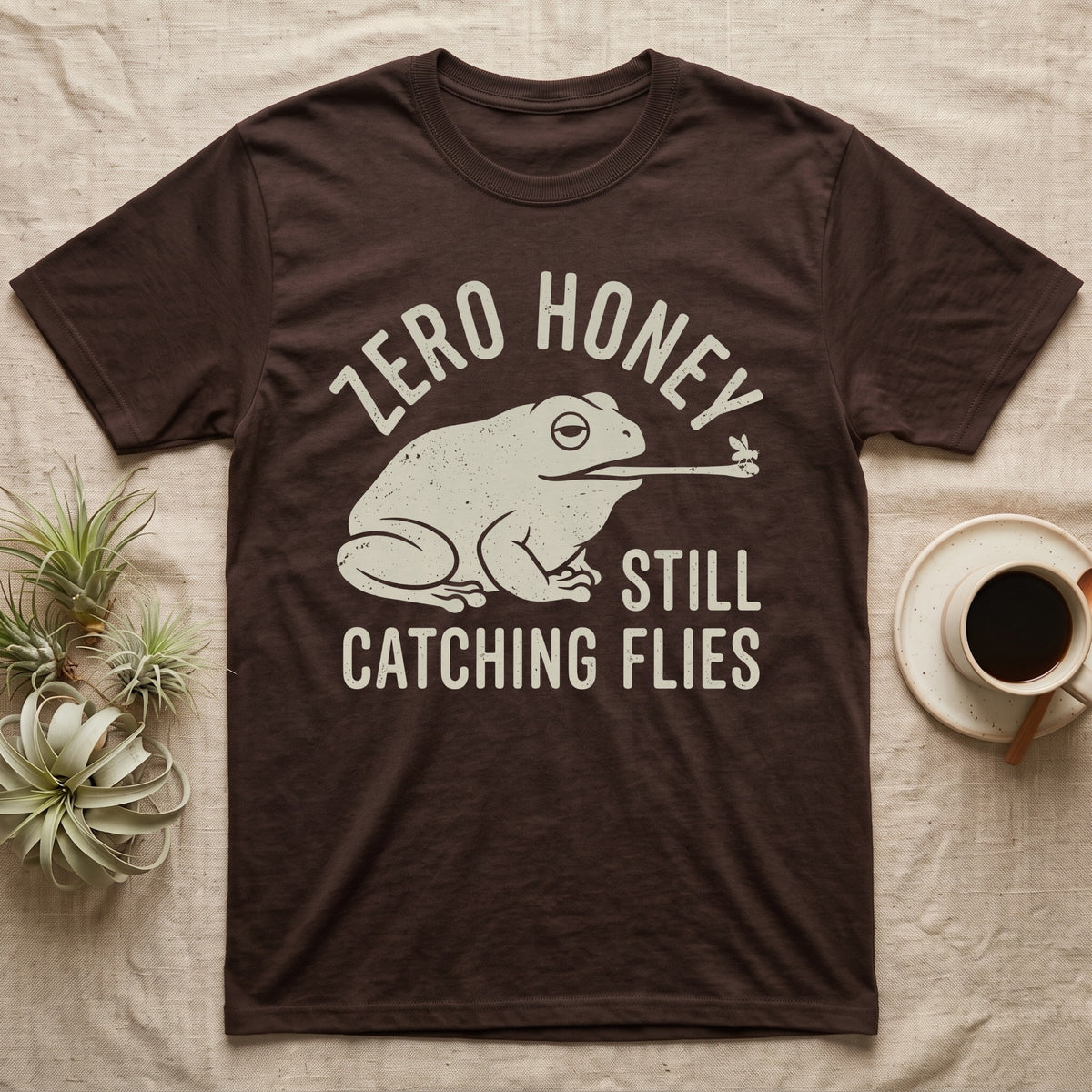 Tree Frog Proverb T-Shirt in Dark Chocolate — Worn cream print of a dumpy White's tree frog catching a fly with its tongue under the words Zero Honey, Still Catching Flies