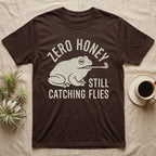 Tree Frog Proverb T-Shirt in Dark Chocolate — Worn cream print of a dumpy White's tree frog catching a fly with its tongue under the words Zero Honey, Still Catching Flies