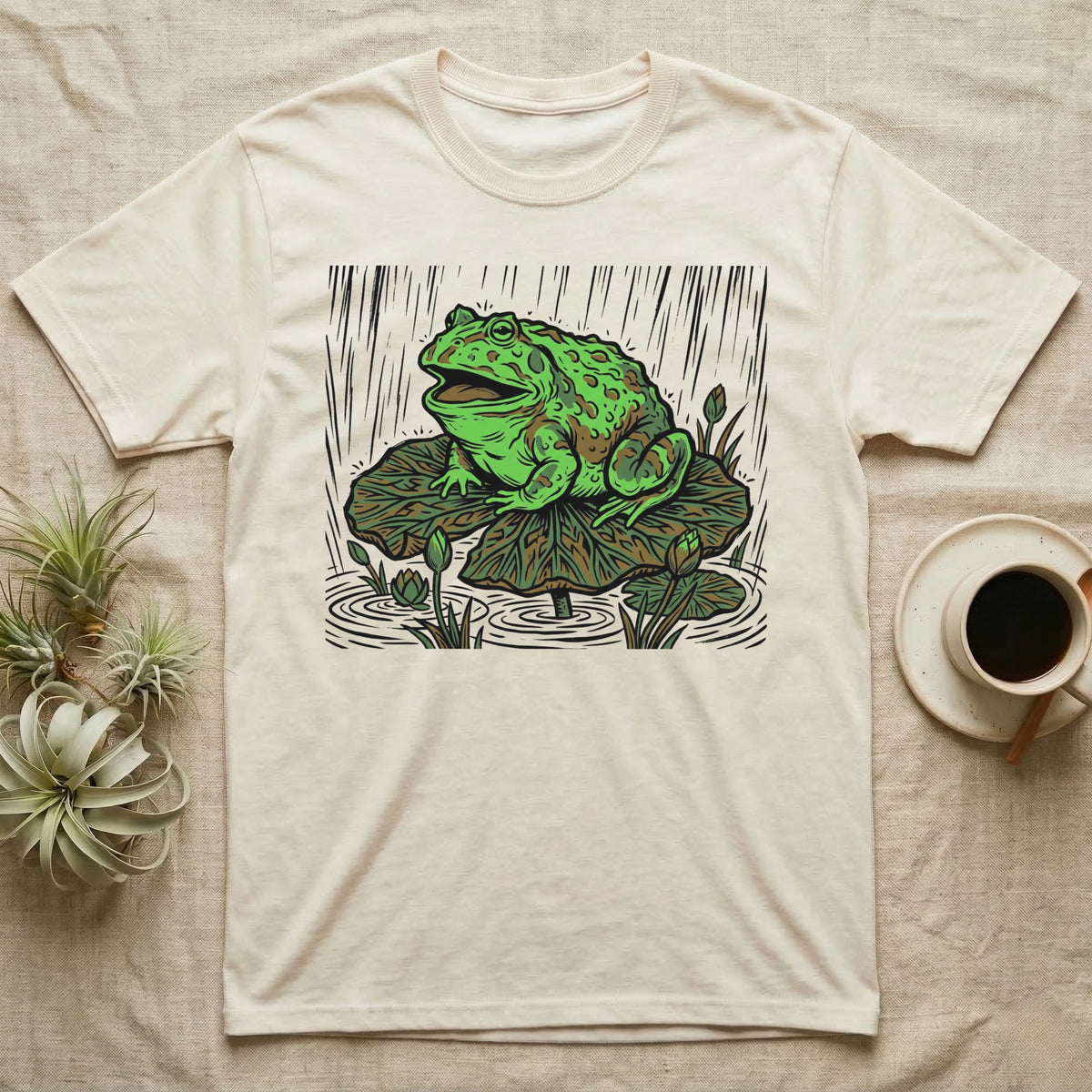 Green frog on a lily pad with rain falling, printed on a cream t-shirt.