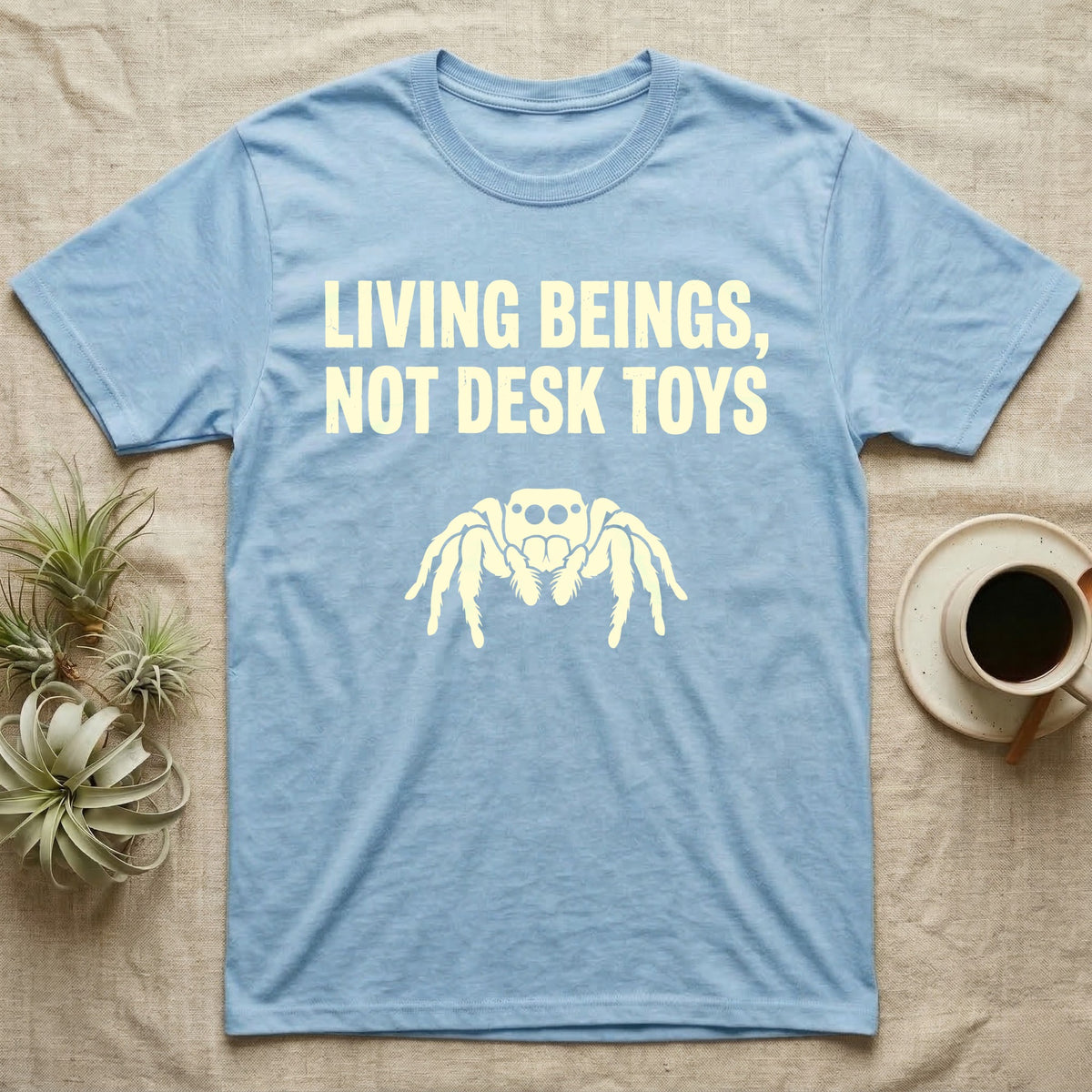 Living Beings Spider T-Shirt in Light Blue — Bold typographic broadside reading Living Beings Not Desk Toys above a small front-facing jumping spider silhouette.