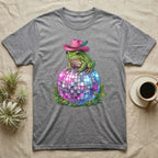 Frog wearing a pink cowboy hat sits on a disco ball with ferns.