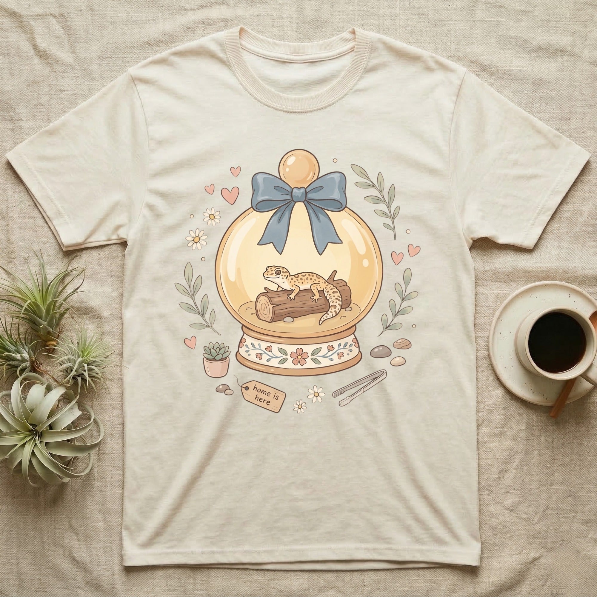 T-shirt featuring a whimsical snow globe with a gecko and "home is here" tag.