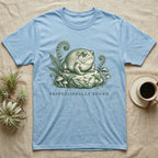 Light blue t-shirt with a cartoon frog sitting on a rock, surrounded by ferns and mushrooms.