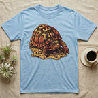 Vintage ink illustration of an Eastern box turtle crawling across sandy pebbles, high-domed shell shown in rust-red and amber, no on-shirt text. — shown on a Light Blue tee.