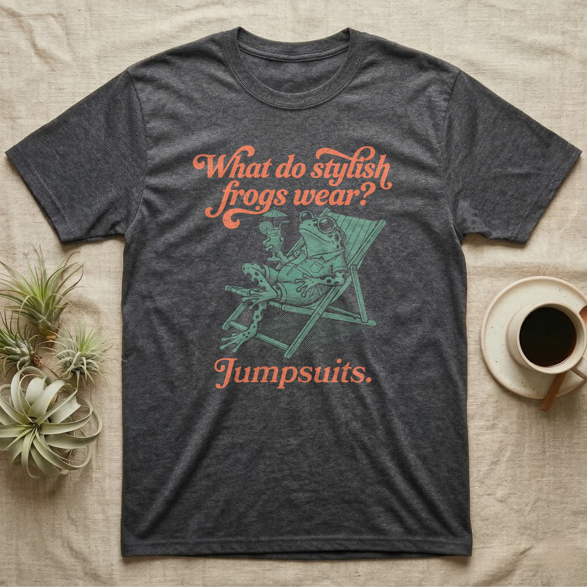 Poison Dart Frog Jumpsuit T-Shirt in Dark Heather — Illustrated green and black poison dart frog reclining in a beach chair with sunglasses and a cocktail, coral script asking what stylish frogs wear, answer jumpsuits.