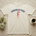 Crested Gecko Hanging T-Shirt in Natural — Red crested gecko hanging by its front feet from the navy arched words HANGING IN THERE