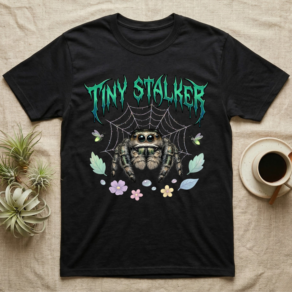 Regal Jumping Spider Feral T-Shirt in Black — Fuzzy jumping spider with huge eyes in a pastel garden under a spiky iridescent-teal metal wordmark reading TINY STALKER.