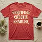 Certified Crestie Enabler T-Shirt in Heather Red — Bold retro drop-shadow wordmark reading "Certified Crestie Enabler" in cream and ochre on a grey background.