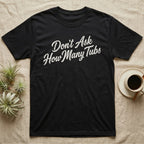 Snake Tubs T-Shirt in Black — Worn off-white 1970s retro brush script climbing a diagonal on charcoal, reading Don't Ask How Many Tubs