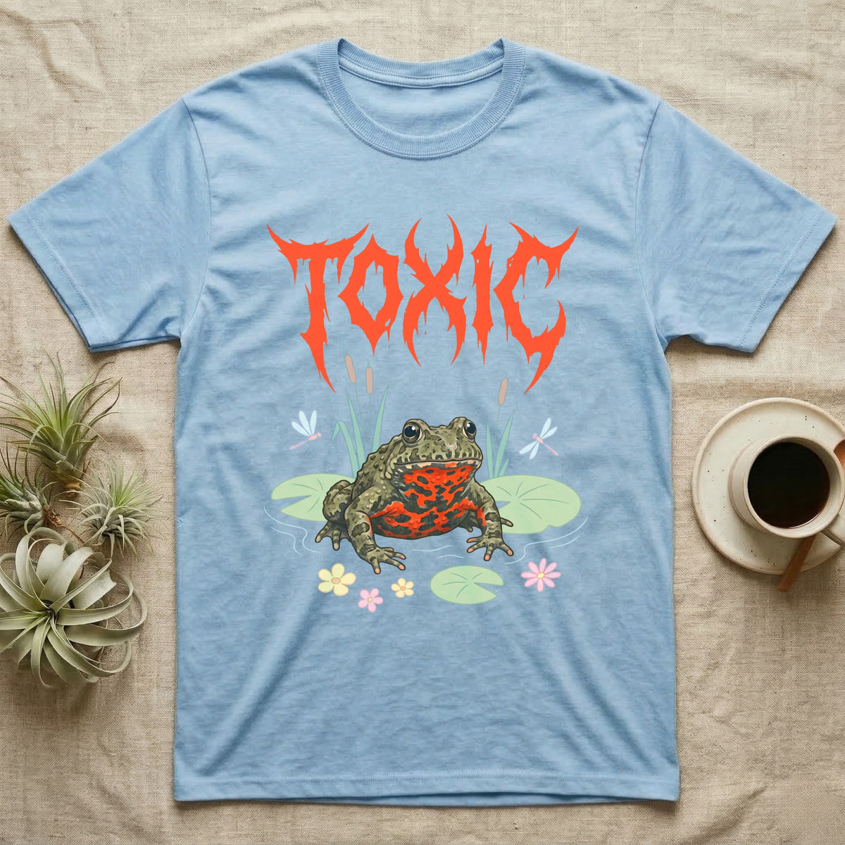 Oriental Fire-Bellied Toad Feral T-Shirt in Light Blue — Pastel fire-bellied toad with red-orange belly among lily pads and dragonflies, jagged metal wordmark "TOXIC" above.