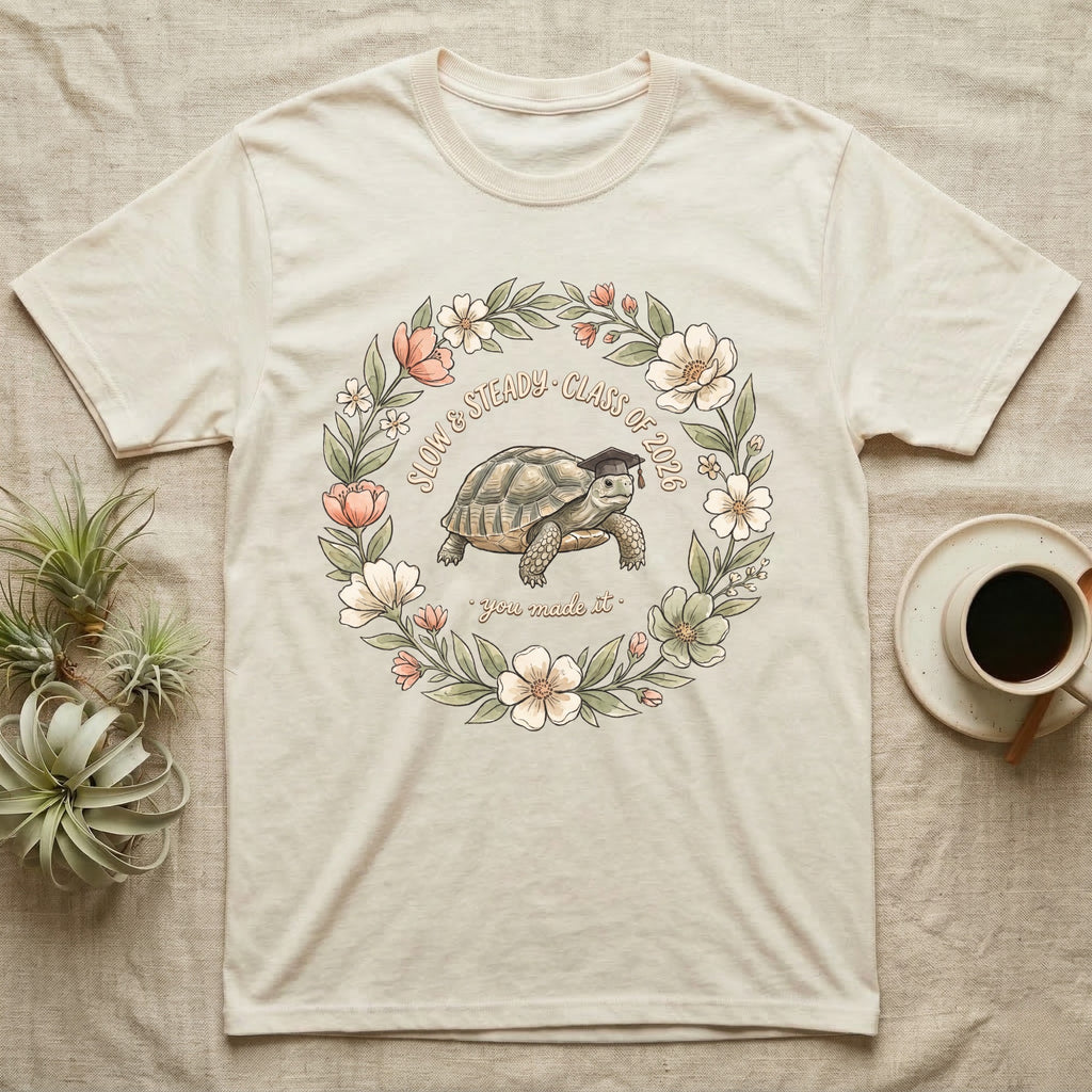 Slow And Steady Graduation T-Shirt in Natural — Watercolour illustration of a tortoise in a graduation cap inside a floral wreath with "Slow & Steady, Class of 2026."