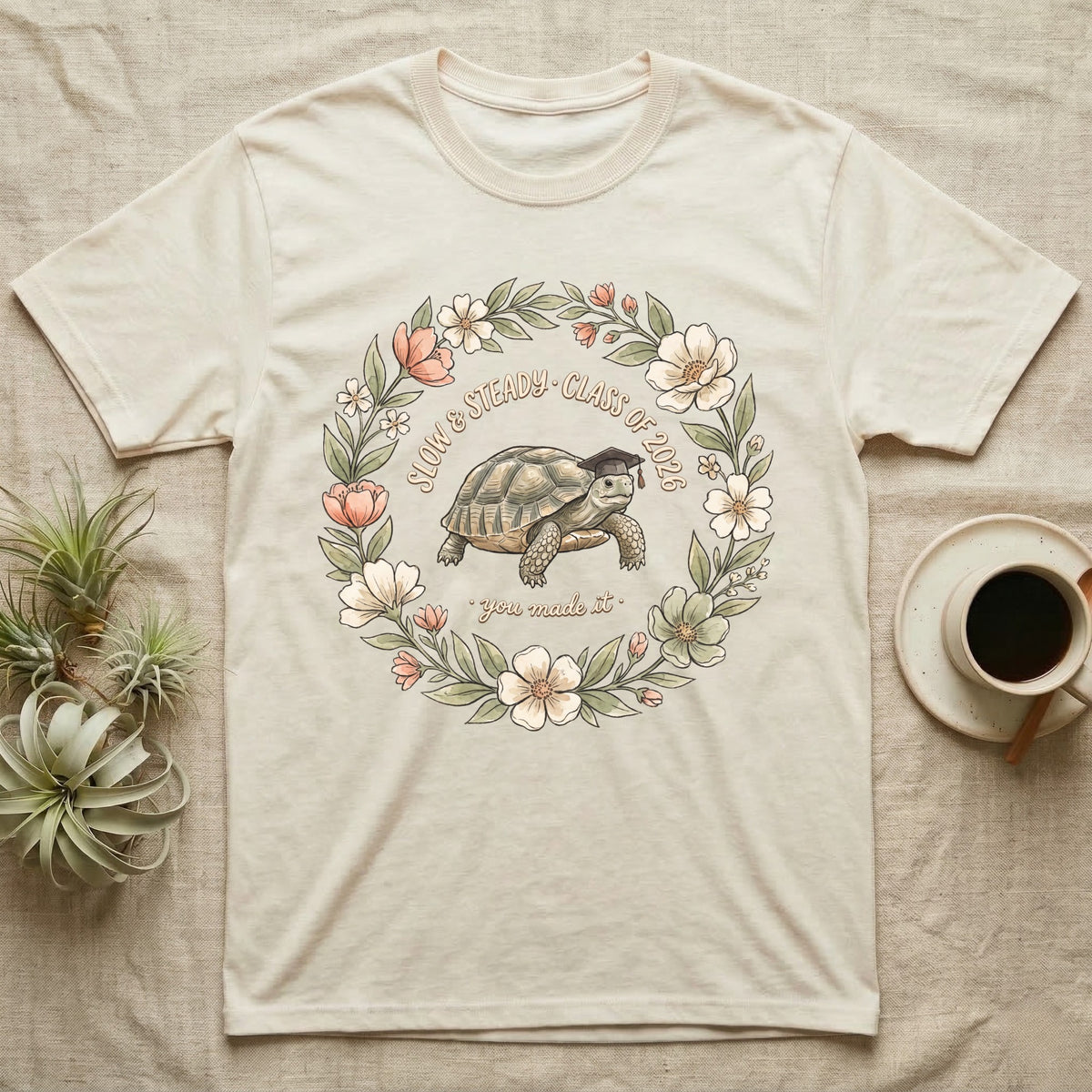 Slow And Steady Graduation T-Shirt in Natural — Watercolour illustration of a tortoise in a graduation cap inside a floral wreath with "Slow & Steady, Class of 2026."