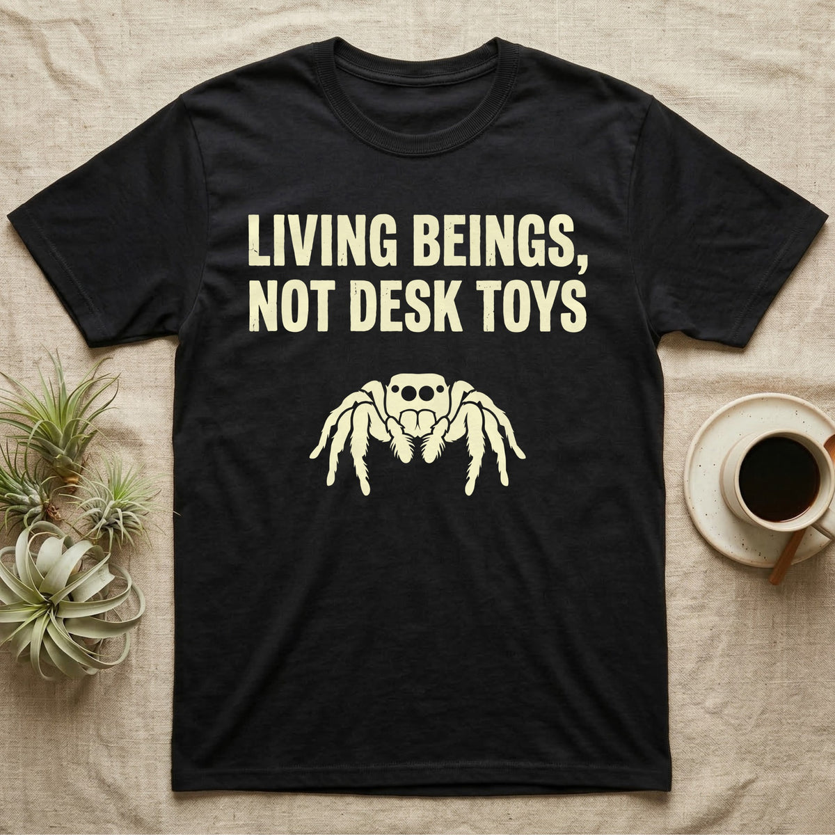 Living Beings Spider T-Shirt in Black — Bold typographic broadside reading Living Beings Not Desk Toys above a small front-facing jumping spider silhouette.