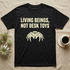 Living Beings Spider T-Shirt in Black — Bold typographic broadside reading Living Beings Not Desk Toys above a small front-facing jumping spider silhouette.