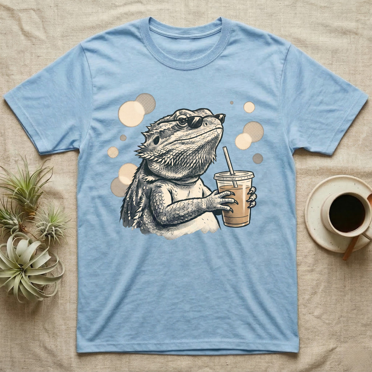 Cold Brew Bearded Dragon T-Shirt in Light Blue — Flat near-photoreal illustration of a bearded dragon in tiny sunglasses holding a cold brew with warm bokeh.