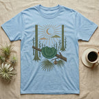 Green Tree Python Home Range T-Shirt in Light Blue — Woodcut-style illustration of a green tree python coiled in a saddle over a branch with palms and tall trunks behind, no on-shirt text.