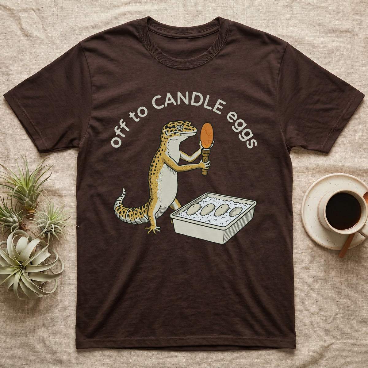 Leopard Gecko Candling T-Shirt in Dark Chocolate — Leopard gecko holding a torch under a glowing orange egg beside a tub of eggs, under the words off to CANDLE eggs