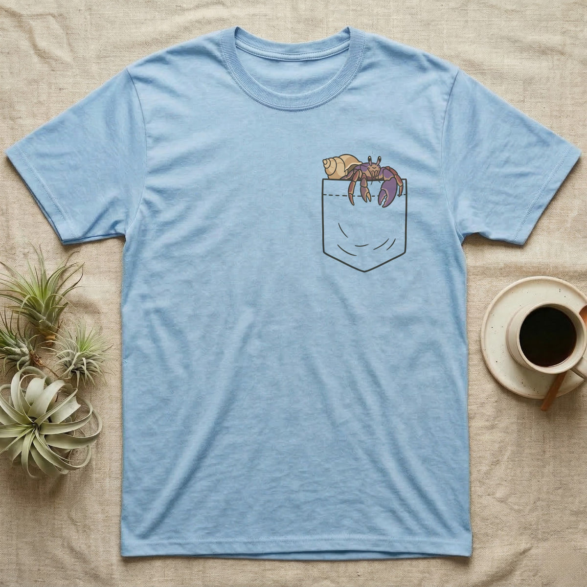 Hermit Crab Pocket T-Shirt in Light Blue — Cartoon purple pincher hermit crab peeking out of a chest pocket, its empty shell on the pocket edge