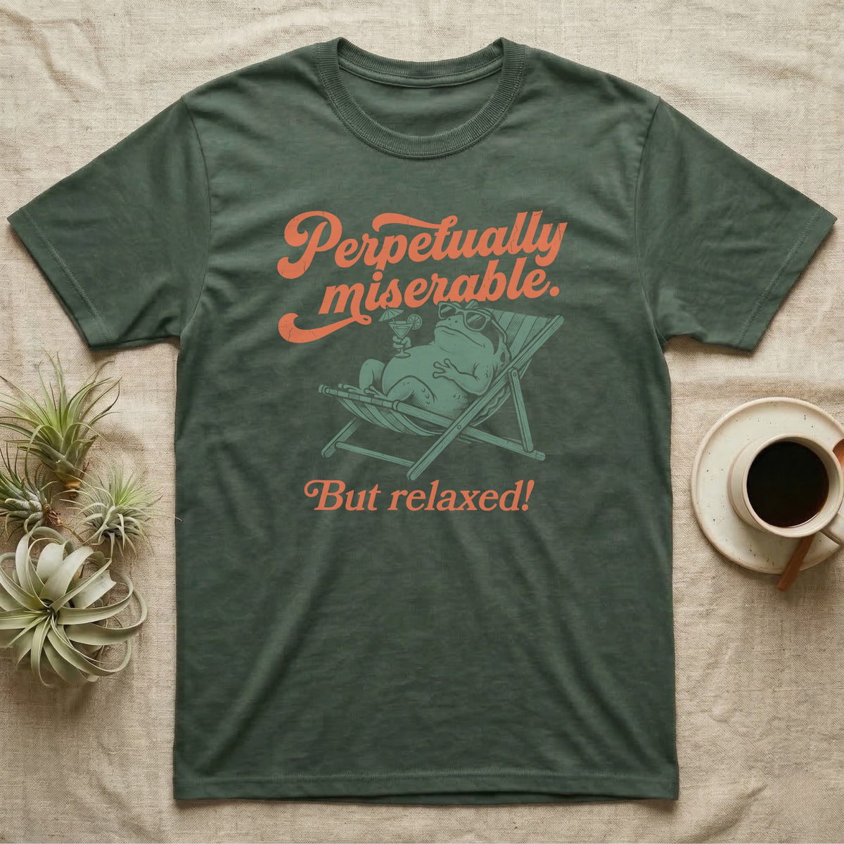 Desert Rain Frog Grump Mode T-Shirt in Military Green — Illustrated desert rain frog lounging in a beach chair with sunglasses and a cocktail, coral script reading Perpetually Miserable But Relaxed.