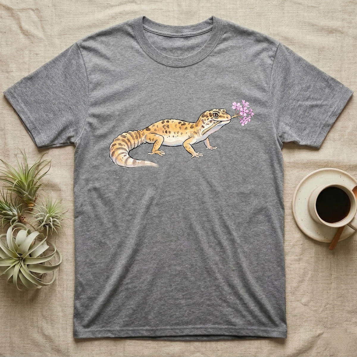 Leopard Gecko Blossom T-Shirt in Sport Grey — Watercolor illustration of a sandy-gold spotted leopard gecko holding a sand verbena blossom sprig, on a dark grey t-shirt.