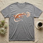Crested Gecko Blossom T-Shirt in Sport Grey — Watercolor illustration of a rust-brown crested gecko holding a frangipani blossom sprig, on a dark grey t-shirt.