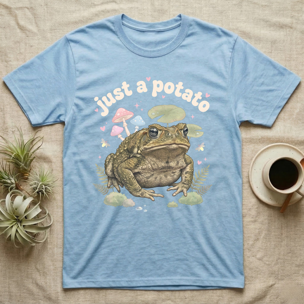 Cane Toad Baby T-Shirt in Light Blue — Warty cane toad among pastel toadstools and lily pads under puffy cream bubble text "just a potato".