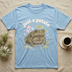 Cane Toad Baby T-Shirt in Light Blue — Warty cane toad among pastel toadstools and lily pads under puffy cream bubble text "just a potato".