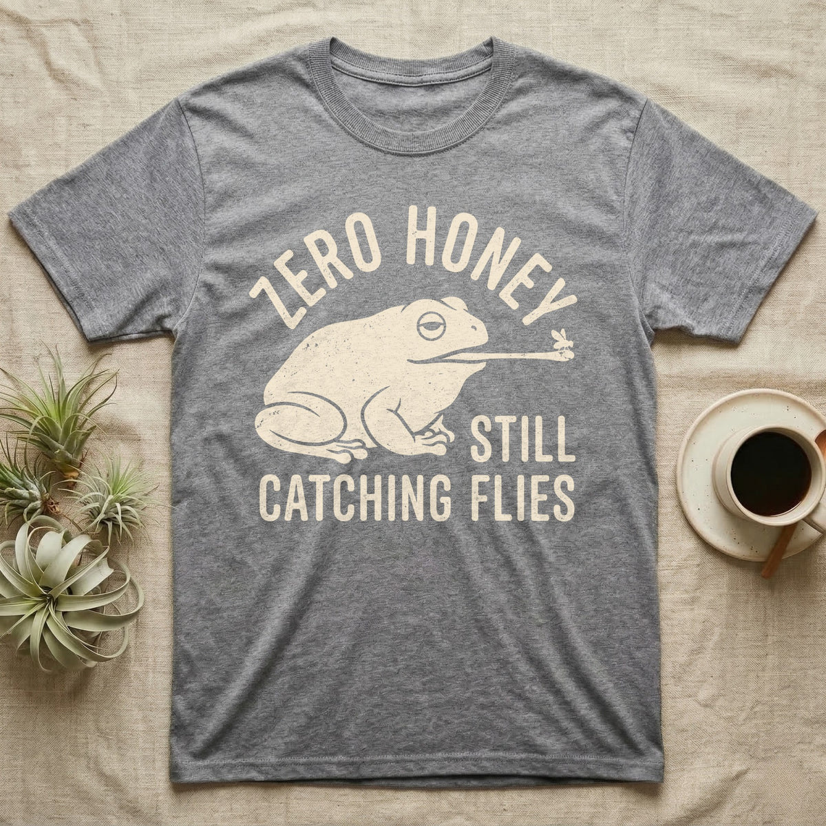 Tree Frog Proverb T-Shirt in Sport Grey — Worn cream print of a dumpy White's tree frog catching a fly with its tongue under the words Zero Honey, Still Catching Flies