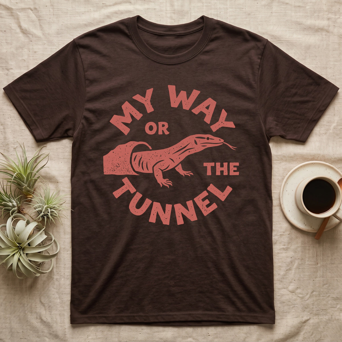 Ackie Monitor Tunnel T-Shirt in Dark Chocolate — Carved one-ink badge of an ackie monitor emerging from its sand tunnel, tongue out, ringed by the words MY WAY OR THE TUNNEL