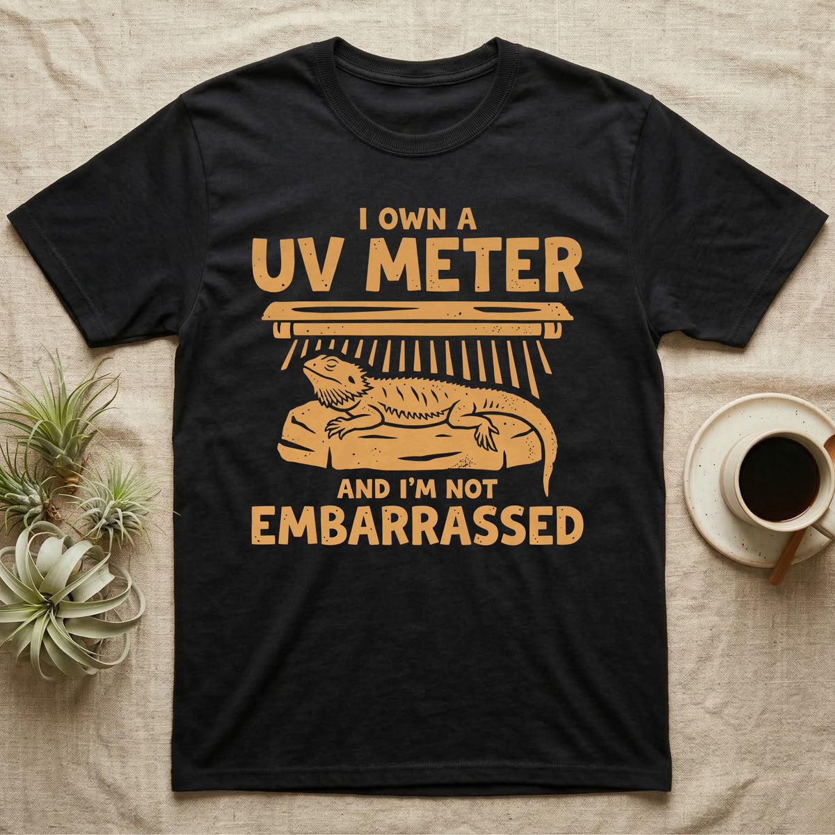 Reptile Keeper UV Meter T-Shirt in Black — Linocut block-print badge of a UVB tube lamp above an empty basking rock, ringed by carved slogan lettering.