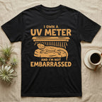 Reptile Keeper UV Meter T-Shirt in Black — Linocut block-print badge of a UVB tube lamp above an empty basking rock, ringed by carved slogan lettering.