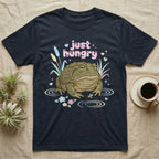 African Bullfrog Baby T-Shirt in Navy — Pastel African bullfrog eyeing a tiny bug among reeds and flowers, puffy pink bubble wordmark "just hungry" above.