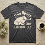 Tree Frog Proverb T-Shirt in Dark Heather — Worn cream print of a dumpy White's tree frog catching a fly with its tongue under the words Zero Honey, Still Catching Flies