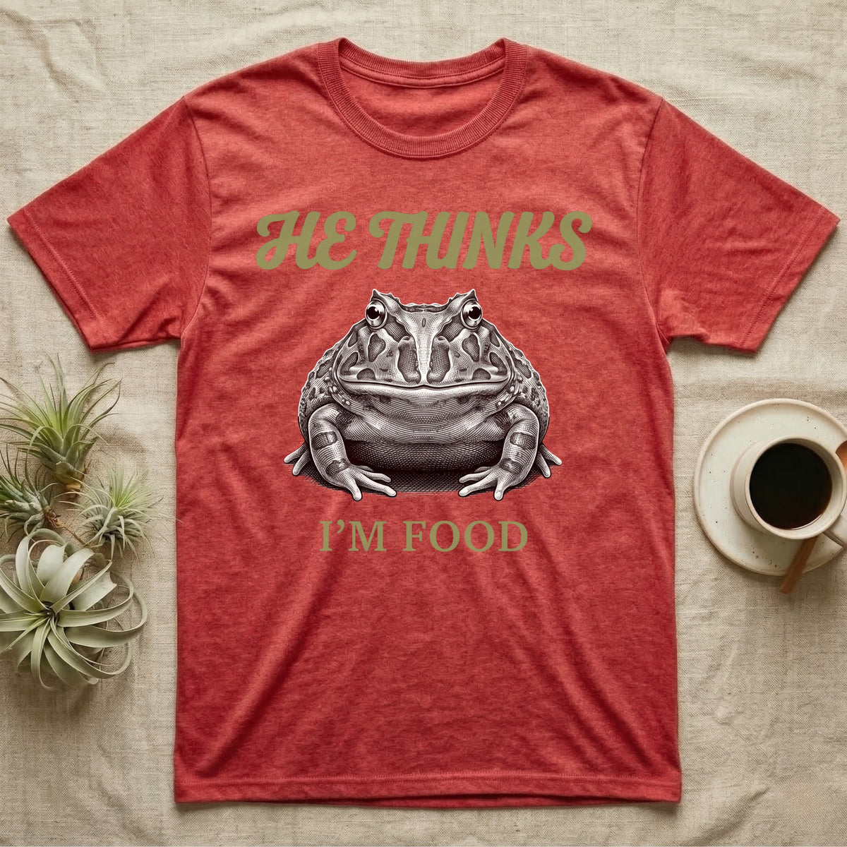 Pacman Frog He Thinks I'm Food T-Shirt in Heather Red — Engraved white-line illustration of a pacman frog staring straight ahead below retro script lettering reading HE THINKS.