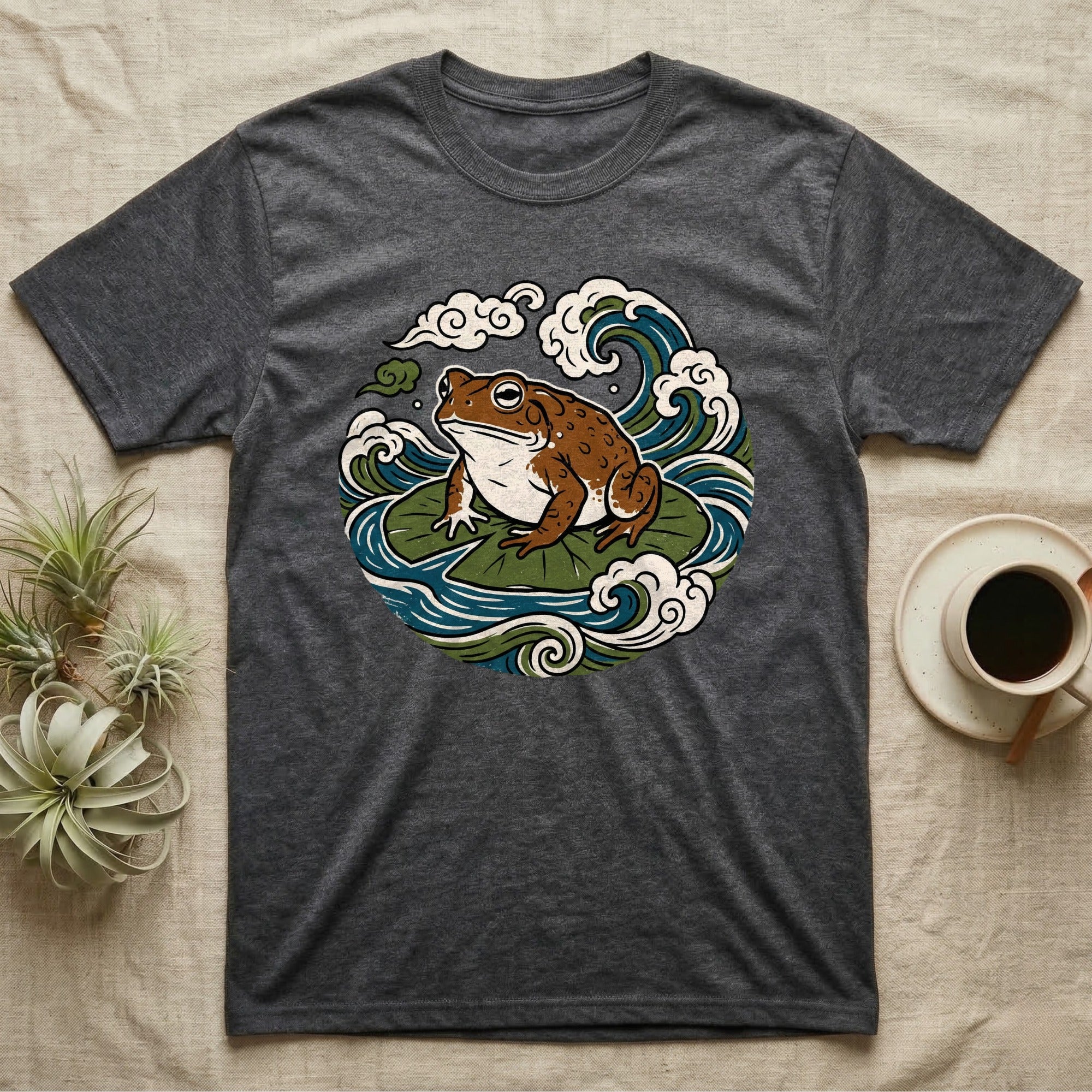 Dark gray t-shirt featuring a stylized frog on lily pads with waves and clouds.