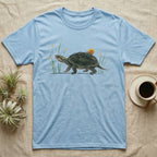 Musk Turtle Hitchhiker T-Shirt in Light Blue — Watercolour musk turtle walking left with a ramshorn snail on its shell, beside slender grass blades with tiny amber flowers.