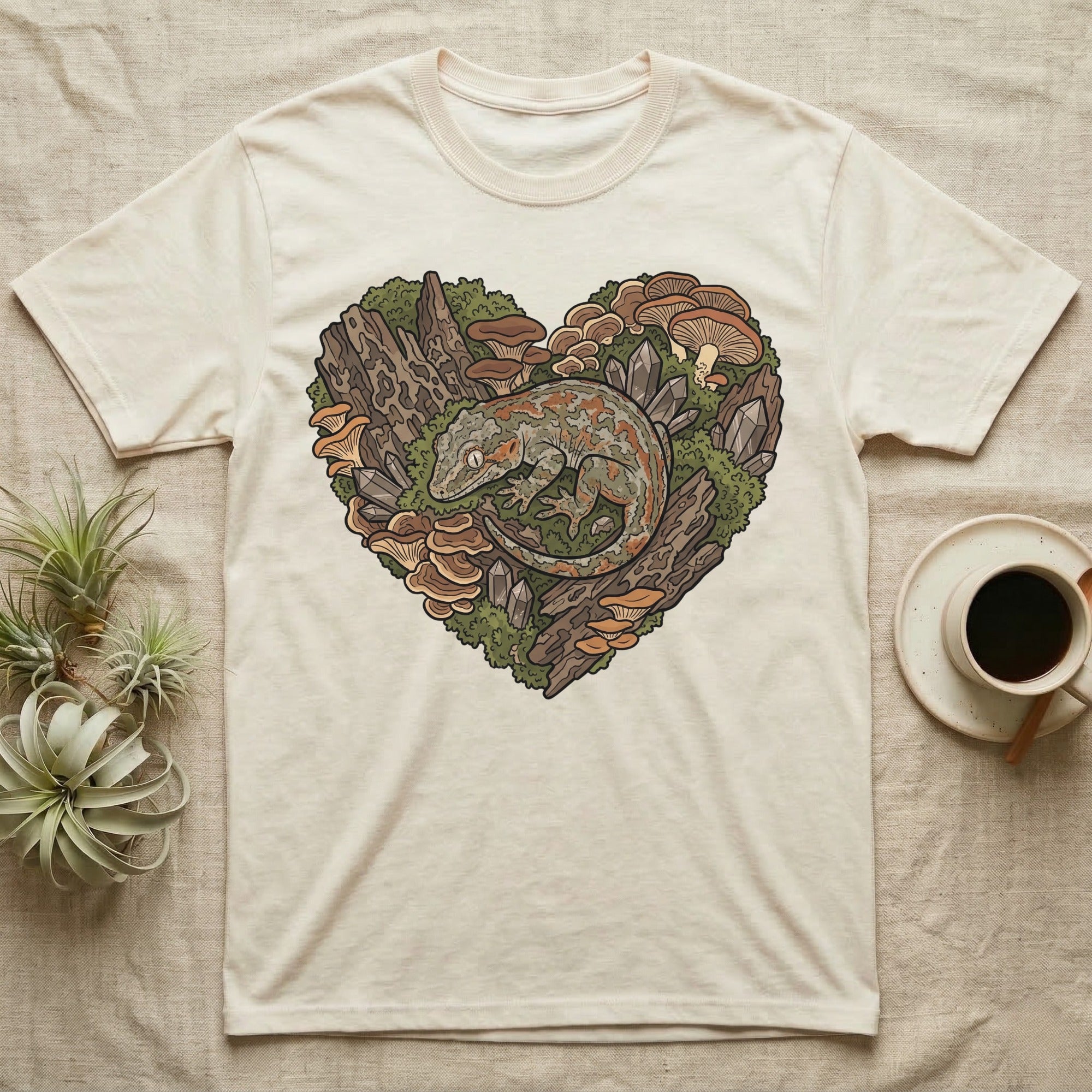 Crested gecko illustration within a heart-shaped forest scene on a cream t-shirt.