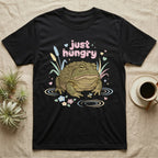 African Bullfrog Baby T-Shirt in Black — Pastel African bullfrog eyeing a tiny bug among reeds and flowers, puffy pink bubble wordmark "just hungry" above.