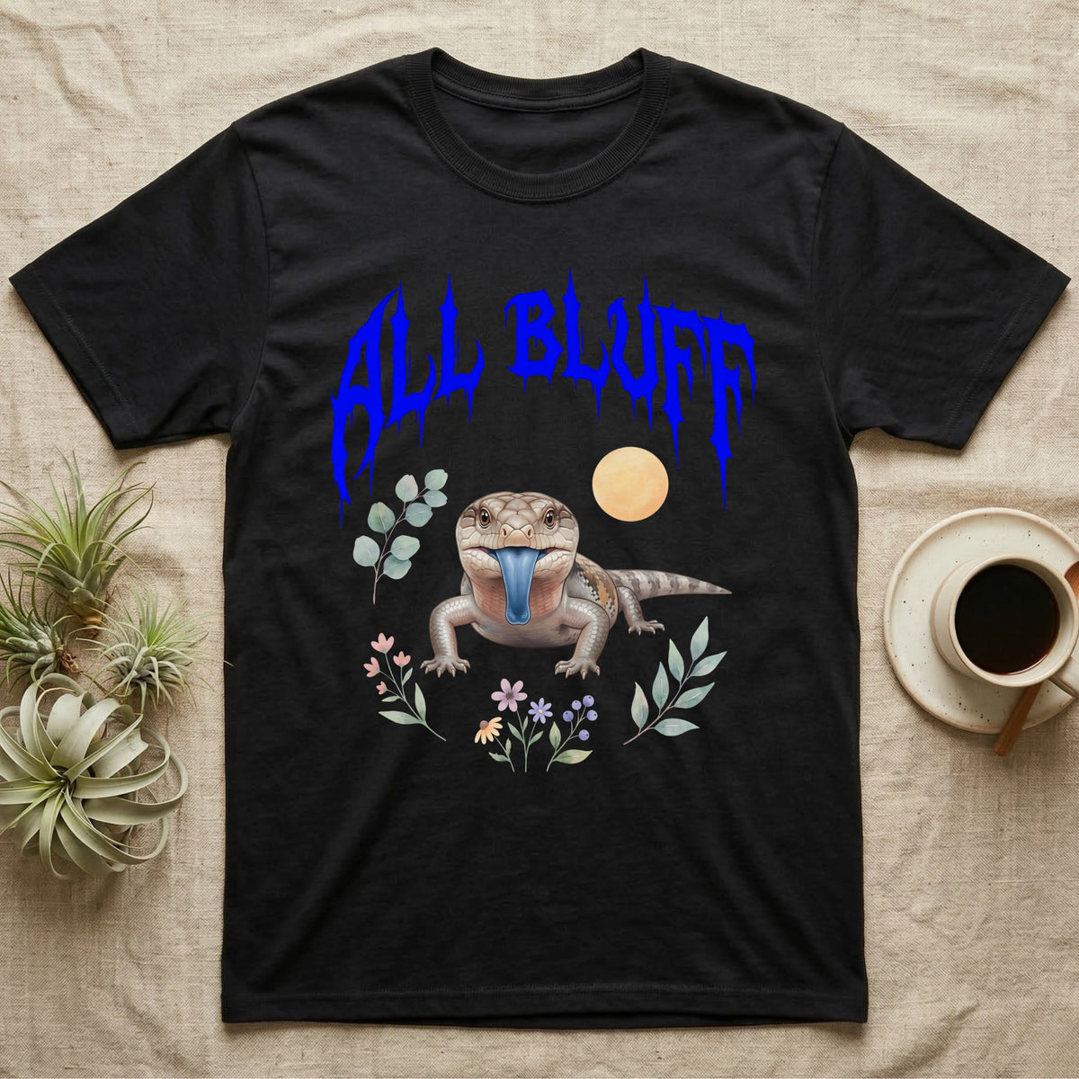Northern Blue-Tongue Skink Feral T-Shirt in Black — Blue-tongue skink with its blue tongue out under a jagged cobalt-blue metal "ALL BLUFF" wordmark and pastel leaves.