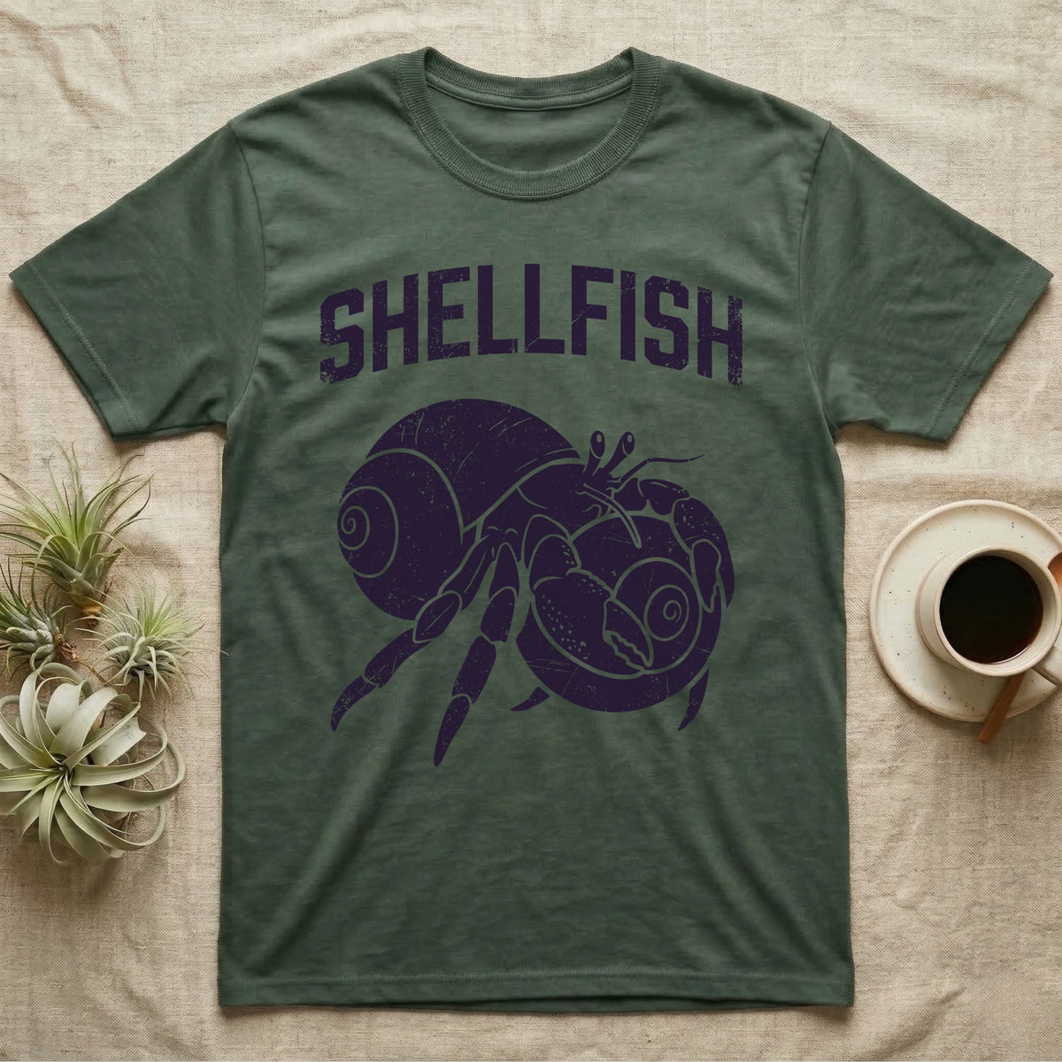 Hermit Crab Shellfish T-Shirt in Military Green — Worn purple one-ink print of a hermit crab hugging a big spare shell to its chest under the word Shellfish