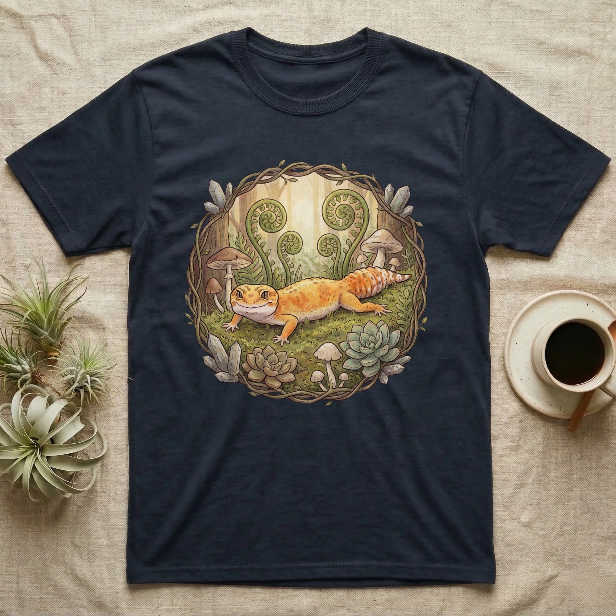 Dark t-shirt featuring a whimsical illustration of a gecko among ferns and mushrooms.