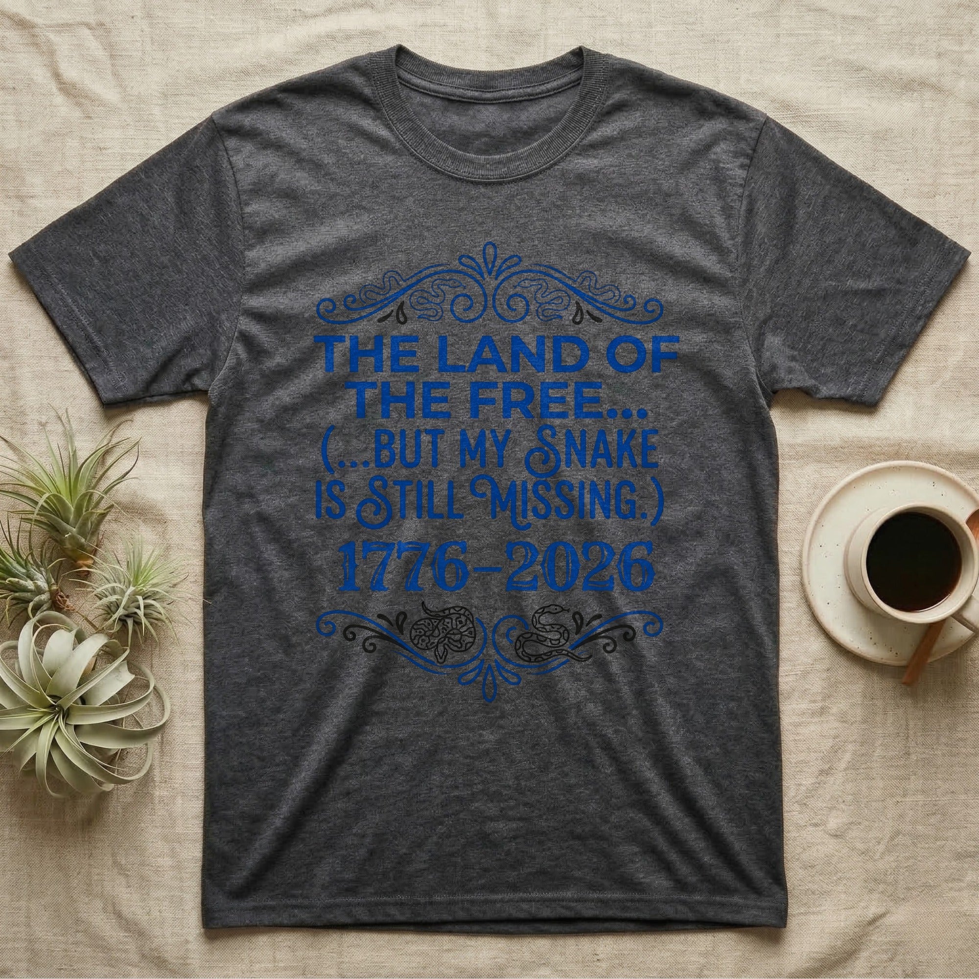 Corn Snake Still Missing Tee in Dark Heather — Royal blue typographic tee reading The land of the free but my snake is still missing, with Victorian filigree and small coiled corn snakes.