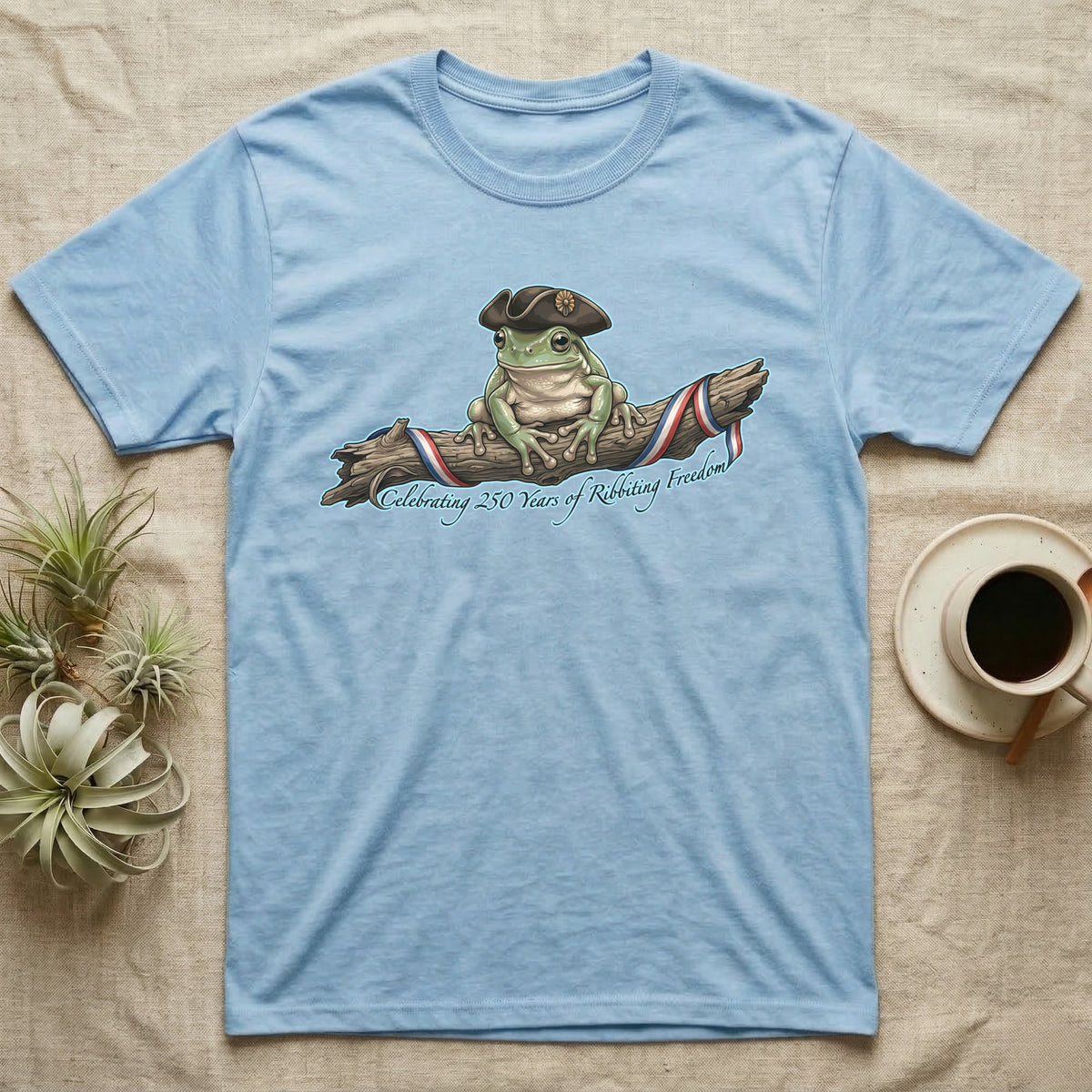 Tree Frog Freedom T-Shirt in Light Blue — Detailed green tree frog in a tricorn hat on a ribboned branch under script Celebrating 250 Years of Freedom.