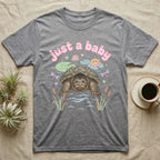 Common Snapping Turtle Baby T-Shirt in Sport Grey — Snapping turtle with hooked beak and spiky shell among pastel lily pads under puffy pink bubble text "just a baby".