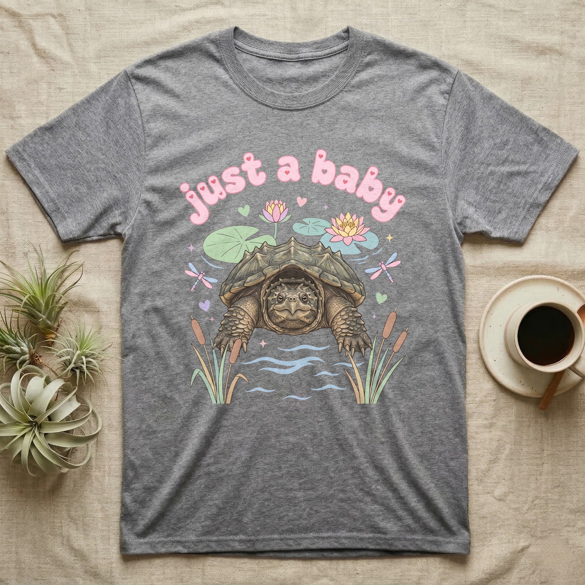 Common Snapping Turtle Baby T-Shirt in Sport Grey — Snapping turtle with hooked beak and spiky shell among pastel lily pads under puffy pink bubble text "just a baby".