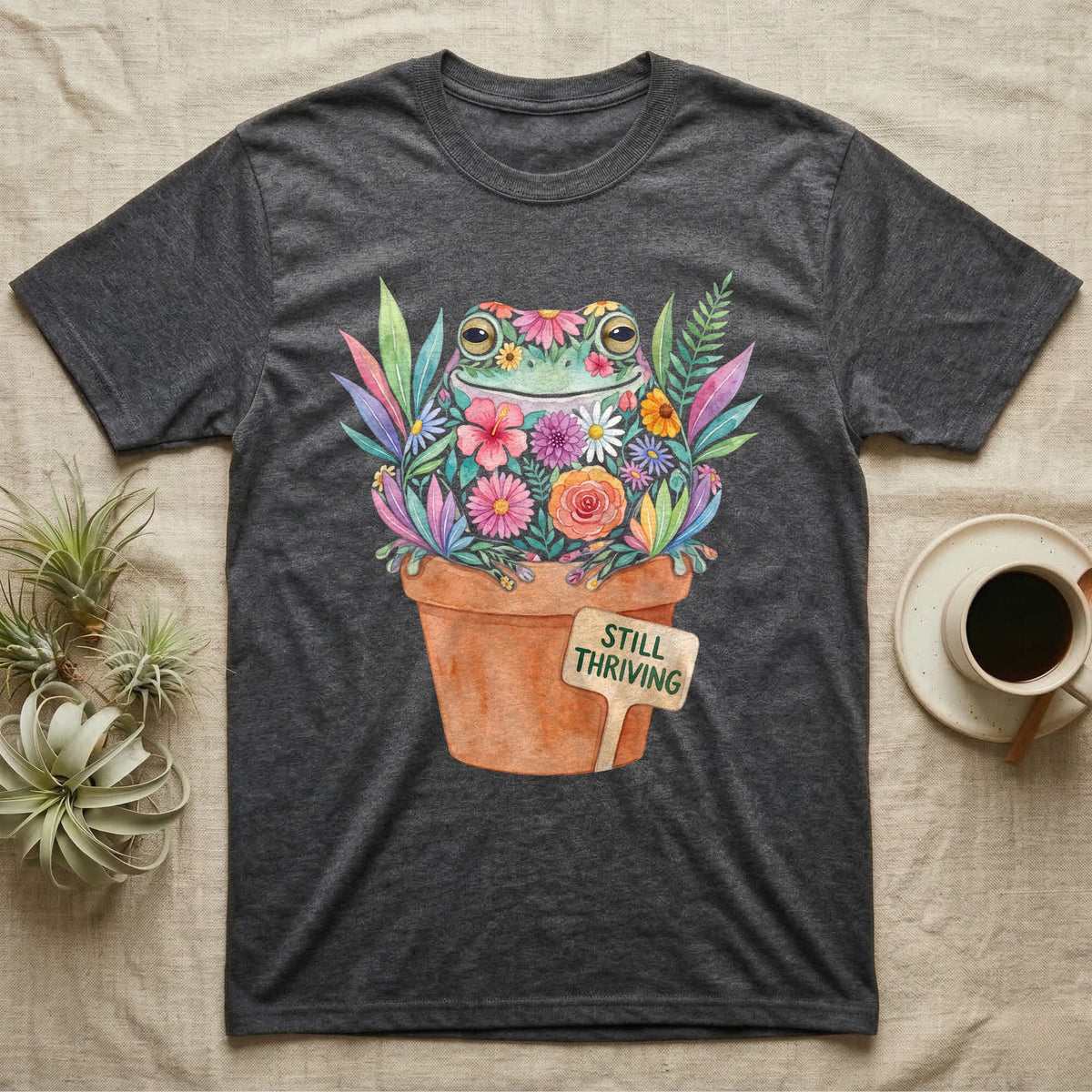 Tree Frog Thriving T-Shirt in Dark Heather — A White's tree frog made of watercolour flowers, sitting in a terracotta pot with a plant label reading STILL THRIVING.